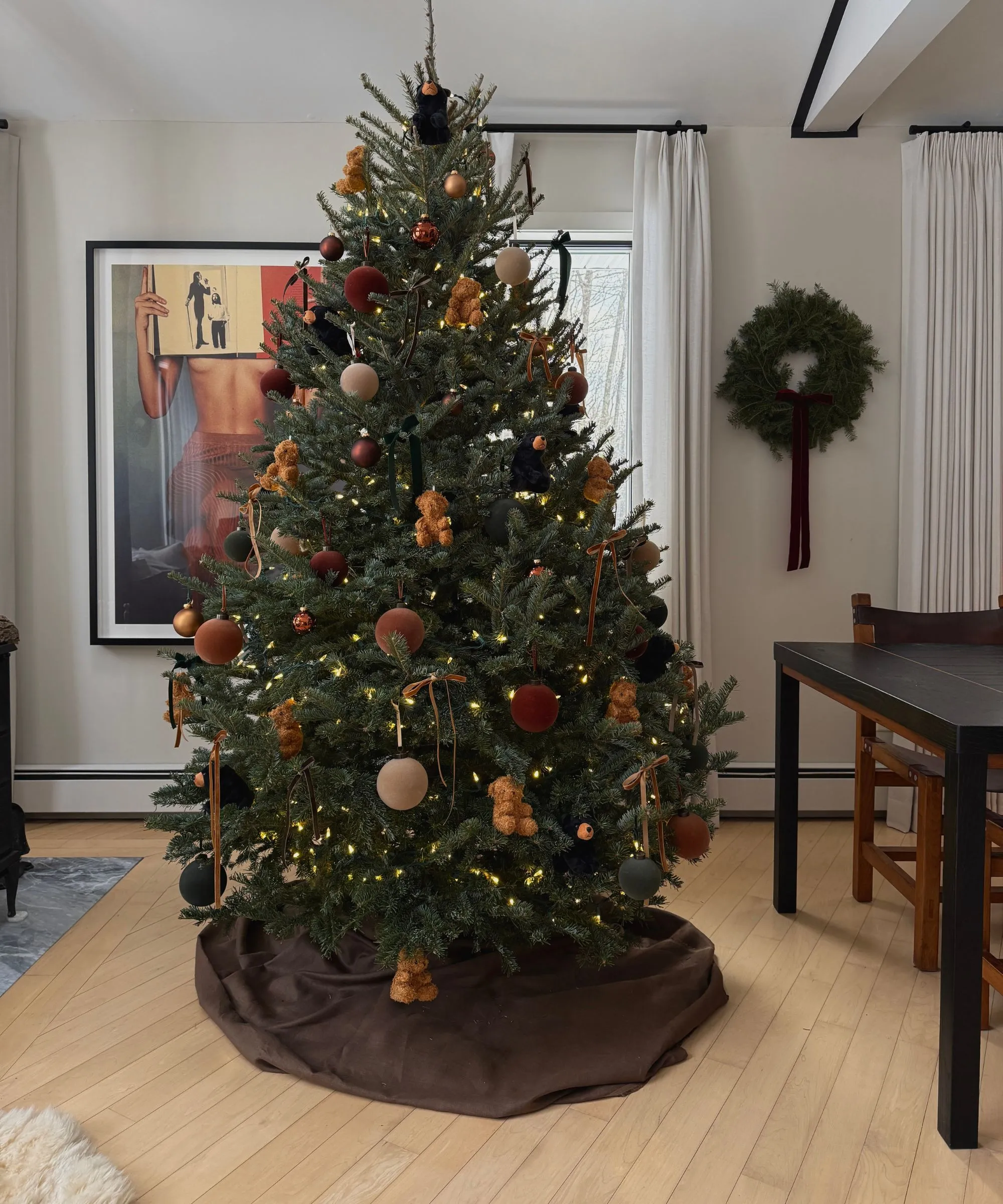 חדר אוכל ניטרלי עם רצפת עץ בלונדינית מעוטרת לחג המולד עם עץ עם חצאית עץ חומה מעוטרת בכדורי קטיפה ודובוני מיני' loading='lazy' title=