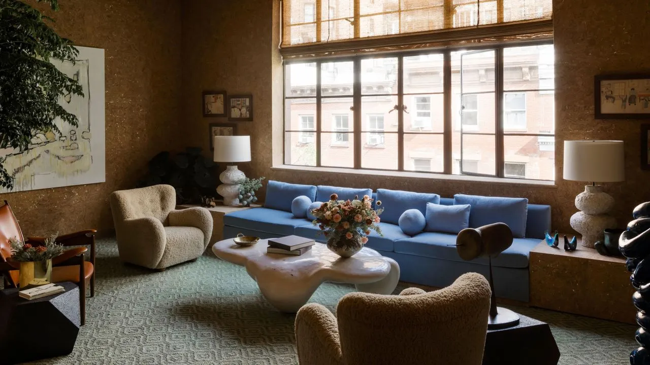 Solbelyst NYC-stue med en overdimensjonert kornblomstblå sofa og khaki lenestoler sammen med et grønt mønstret teppe og korkdekkede vegger.' loading='lazy' title=