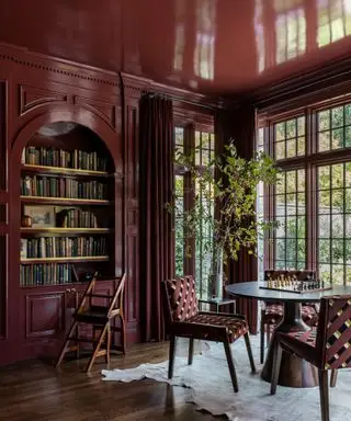 Sala de biblioteca de color rojo oscuro con mesa redonda y ventana grande.