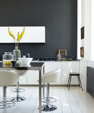 Espacio de cocina con paredes pintadas de grafito, suelos y armarios de cocina blancos, isla de cocina alta en metal y negro, taburetes altos en blanco y metal, jarrones decorativos y adornos sobre la mesa, taburete bajo junto al radiador gris, marcos decorativos y cuadros en la pared y descansando en la encimera