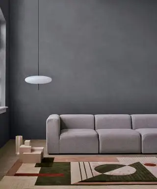 Sala de estar con paredes pintadas de grafito moteado, un gran sofá gris, una alfombra geométrica en colores apagados, un objeto decorativo de madera en el piso, un colgante bajo con una pantalla redondeada de vidrio ópalo