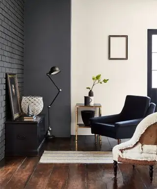 Sala de estar con pared de ladrillo pintado de grafito en el techo, pared pintada de color crema a la derecha, piso de madera oscura, sillón en azul y sillón tapizado en color crema, mesa baja negra con pintura y cuenco decorativo, lámpara de pie negra, mesa auxiliar de metal con jarrones. , pequeña alfombra estampada a rayas