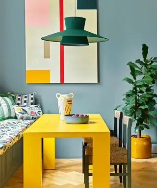 Esszimmer mit blau gestrichenen Wänden, farbenfrohen, abstrakten Kunstwerken an den Wänden, große Pflanze in gelbem Keramiktopf auf dem Boden, leuchtend gelber rechteckiger Esstisch, eingebaute Bank am Fenster, gepolsterte Sitzfläche und Rückenlehne mit Kissen, Tisch mit bunter Vase und Schale dekoriert