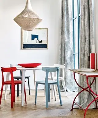 Weiß gestrichenes Esszimmer mit hellem Fischgrätenholzboden, abgerundeter weißer Esstisch mit schwarzen, roten, blauen und weißen Esszimmerstühlen, strukturierter, gewebter Anhänger über dem Tisch, Konsolentisch mit rot lackiertem Metallgestell, Holztischplatte, hellgrau gemusterte Vorhänge auf beiden Seiten helles Fenster, blaue und weiße Kunstwerke an der Wand