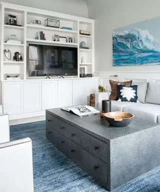 Lys hvit stue med hvit oppbevaring og utstillingsenhet med tv, stort grått salongbord med trekk, blått teppe og blått kunstverk på veggen med bølgedesign