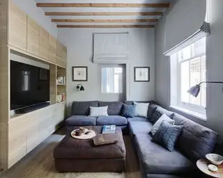 Stue med stor grå L-formet sofa, ottomansk og innebygde skap med TV, trebjelker, gråmalte vegger og persienner, vegglamper og innrammede kunstverk, teksturerte teppe på tregulv
