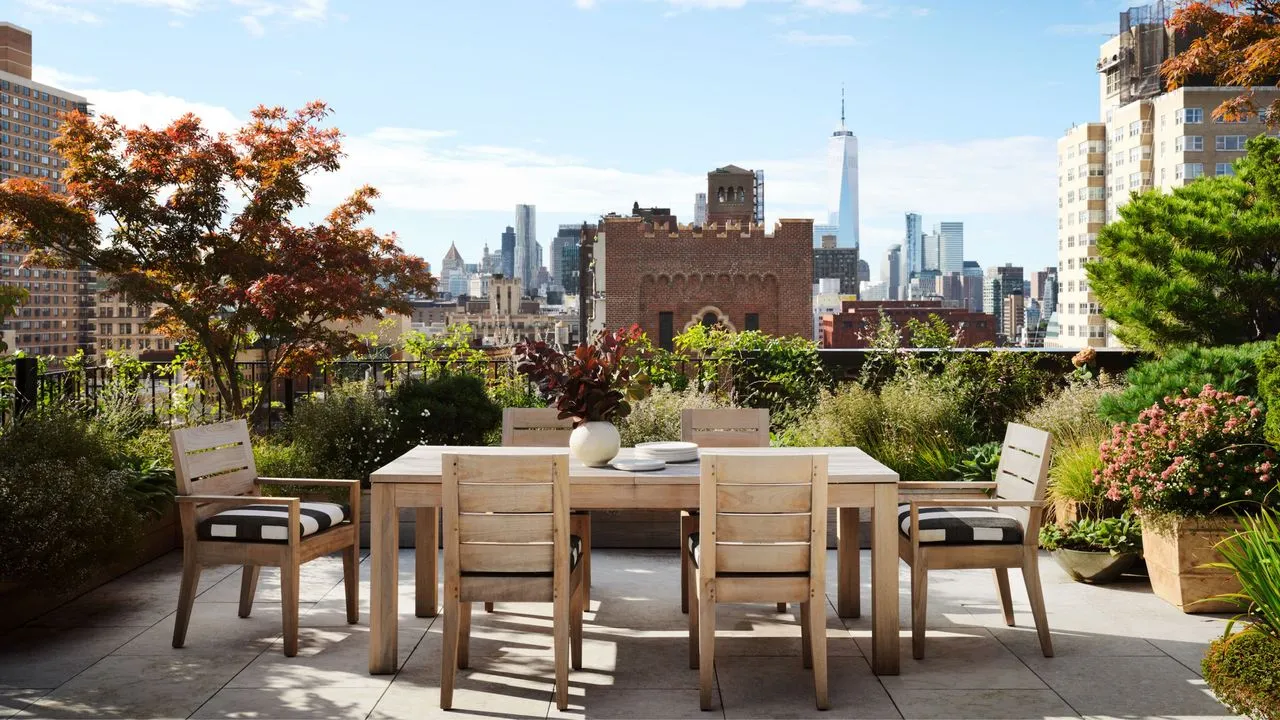 En tagterrasse med udsigt over New Yorks skyline. Et træbord og stole sidder i midten med moden beplantning omkring' loading='lazy' title=