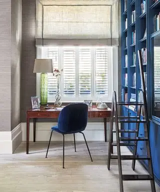 Arbeitszimmer mit Schreibtisch neben dem Fenster und Einbauschränken und dunkelblauen Bücherregalen.