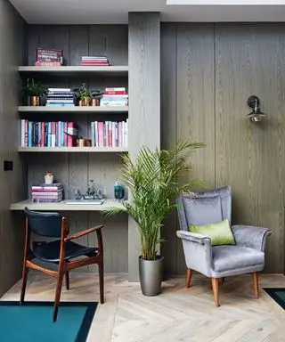 Ecke eines Wohnraums mit eingebautem Schreibtisch und Regal für Bücher, braungraue Holzwände, Schreibtischstuhl aus dunklem Holz und schwarzem Leder, gepolsterter grauer Sessel mit grünem Kissen und Holzbeinen, Metallwandleuchte über dem Sessel, grüne Graspflanze in grauer Pflanze Topf