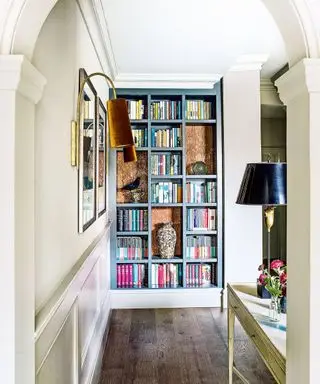 Flur mit cremefarbenen Wänden, dunklem Holzboden, weiß gestrichenem Konsolentisch aus Holz mit schwarzer und metallischer Tischlampe, Blick auf ein eingebautes, blau gestrichenes Bücherregal voller Bücher und Deko, an der Wand zwei gerahmte Bilder mit Wandlampen daneben