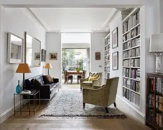 Heller offener Wohn- und Essbereich, weiß gestrichene Wände, raumhohe Regale voller Bücher, gemütliche Sitzecke mit Sofa und zwei Sesseln, darunter ein weicher grauer Strukturteppich, viele gerahmte Bilder und Gemälde an den Wänden, daneben Lese- und Schreibtischlampen Sitzbereiche, rechteckiger Wandspiegel über dem Sofa