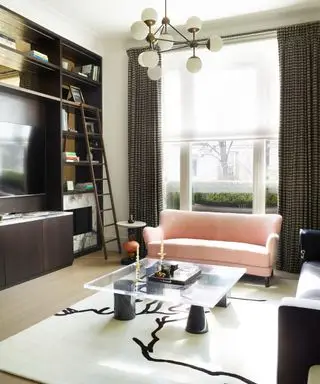 Wohnzimmer mit raumhoher brauner Aufbewahrungs- und Ausstellungseinheit, Holzleiter und Marmorkamin, schwarzem und cremefarbenem Teppich mit quadratischem Couchtisch aus Glas, karierten Vorhängen, schwarzem Sofa und rosa Sofa, buntem Beistelltisch, abgerundeter weißer und metallischer Pendelleuchte