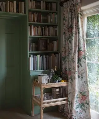 Ecke eines Wohnzimmers im Landhausstil, grün gestrichene Wände und geflochtener, natürlich strukturierter Getränkewagen, großes raumhohes Fenster mit geblümten Vorhängen, großes Bücherregal mit Büchern an der Wand und über der Tür