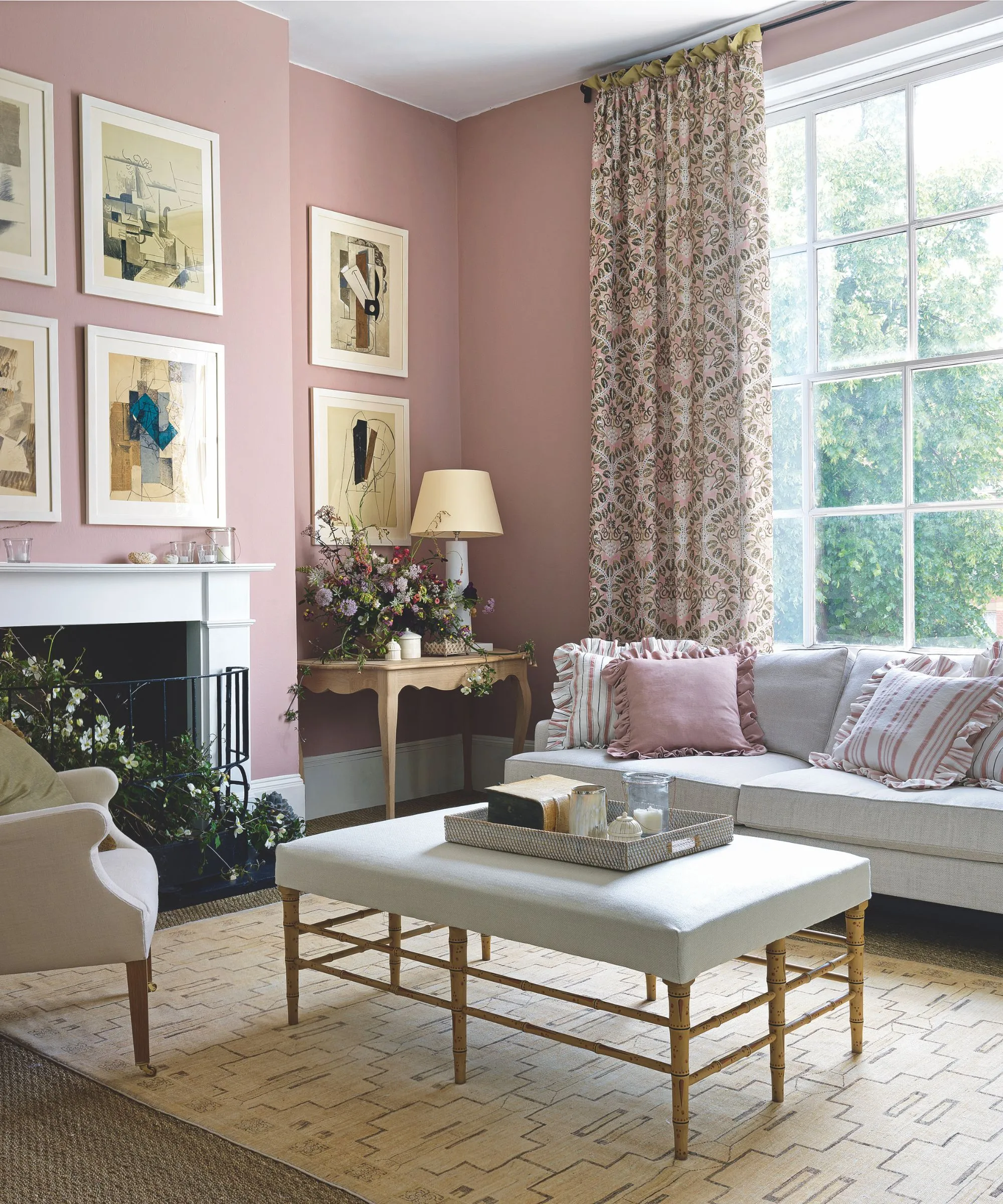 Una sala de estar rosa de estilo vintage con cortinas estampadas frente a una gran ventana con pared de la galería enmarcada blanca, una repisa blanca, un sofá gris con rosa con volantes y cojines a rayas con reposapiés a juego con bandeja y un sillón rosa rubor mirando hacia él' loading='lazy' title=