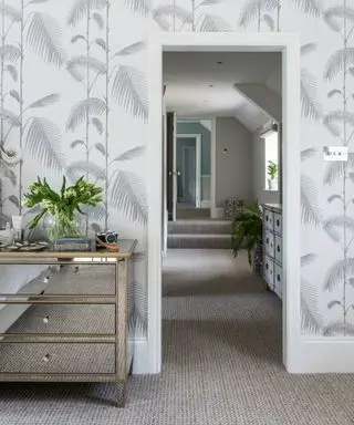 Quarto com carpete, papel de parede com padrão de folhas botânicas cinza claro e branco e cômoda espelhada. Vista ao longo de um corredor no andar superior.