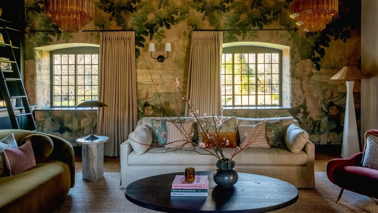 stor stue med botanisk veggmaleri tapet i malerisk stil to store vinduer innebygd til venstre, en nøytral sofa og en fløyelsbuet sofa med et svart avrundet salongbord i tre' loading='lazy' title=