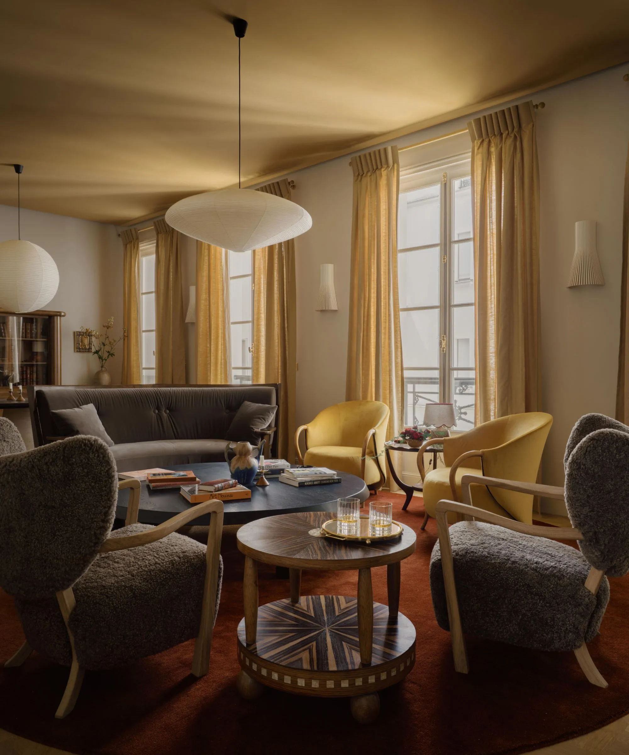 En stue med okerfarget tak varmhvite vegger og stort vindu med okerfargede gardiner. Et mørkerødt teppe og mørke nøytrale stoler.' loading='lazy' title=