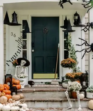 Halloween-Türdekorationsideen mit Fledermausdekorationen an den Wänden, auf dem Verandadach kletternden Spinnen, sitzenden Skeletten, Kürbissen und Hexenhüten, die vom Dach baumeln