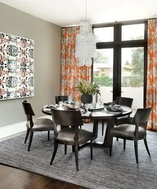 Salle à manger grise avec table et chaises en bois noir, rideaux à motifs orange, œuvres d'art colorées, lustre suspendu au-dessus de la table