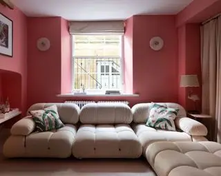 Rosamalt stue med stor kremsofa med vattert design, to frostet glass, avrundede vegglamper og bordlampe med scalloped skjerm, krempersienne og rosa, grønne og hvite puter på sofa