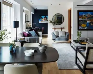 Åpen stue- og familierom, mørk marineblå vegg, mørkt tregulv, soner for sitteplasser, spisestue og arbeid, grå sofa og lenestol, grått teppe og salongbord i hvit marmor, fargerike kunstverk på vegger, avrundet speil, svart spisebord i tre