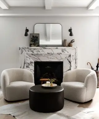 Salon noir et blanc avec deux fauteuils assortis et table basse noire, disposé autour d'une cheminée en marbre, miroir sur cheminée, deux appliques murales assorties, tapis style peau de bête