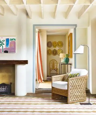 Salon crème avec poutres en bois au plafond, fauteuil en osier, lampadaire élégant, donnant sur le couloir, rideau à rayures, console en bois avec lampe de table, tapis coloré