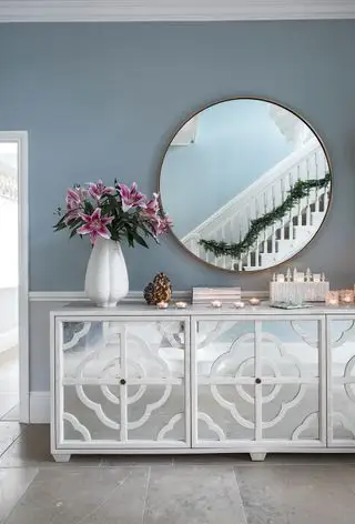 Una credenza a specchio e uno specchio sopra in un corridoio con pareti azzurre con un riflesso di ringhiere con una ghirlanda
