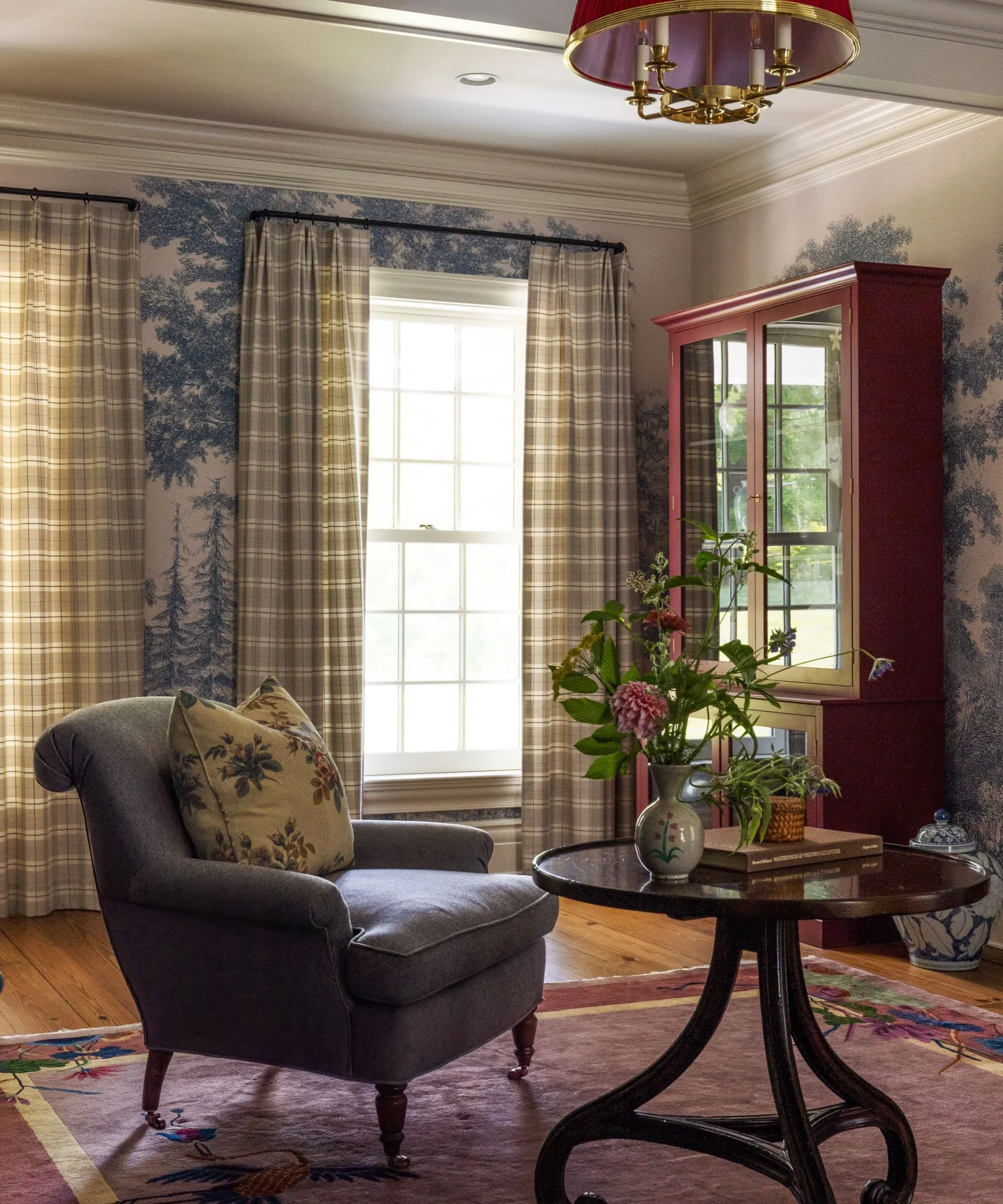 파란색과 흰색 나무 벽화 벽지로 장식된 아늑한 거실 안락의자와 테이블, 깅엄 커튼, 나무 옷장, 꽃병에 꽃' loading='lazy' title=