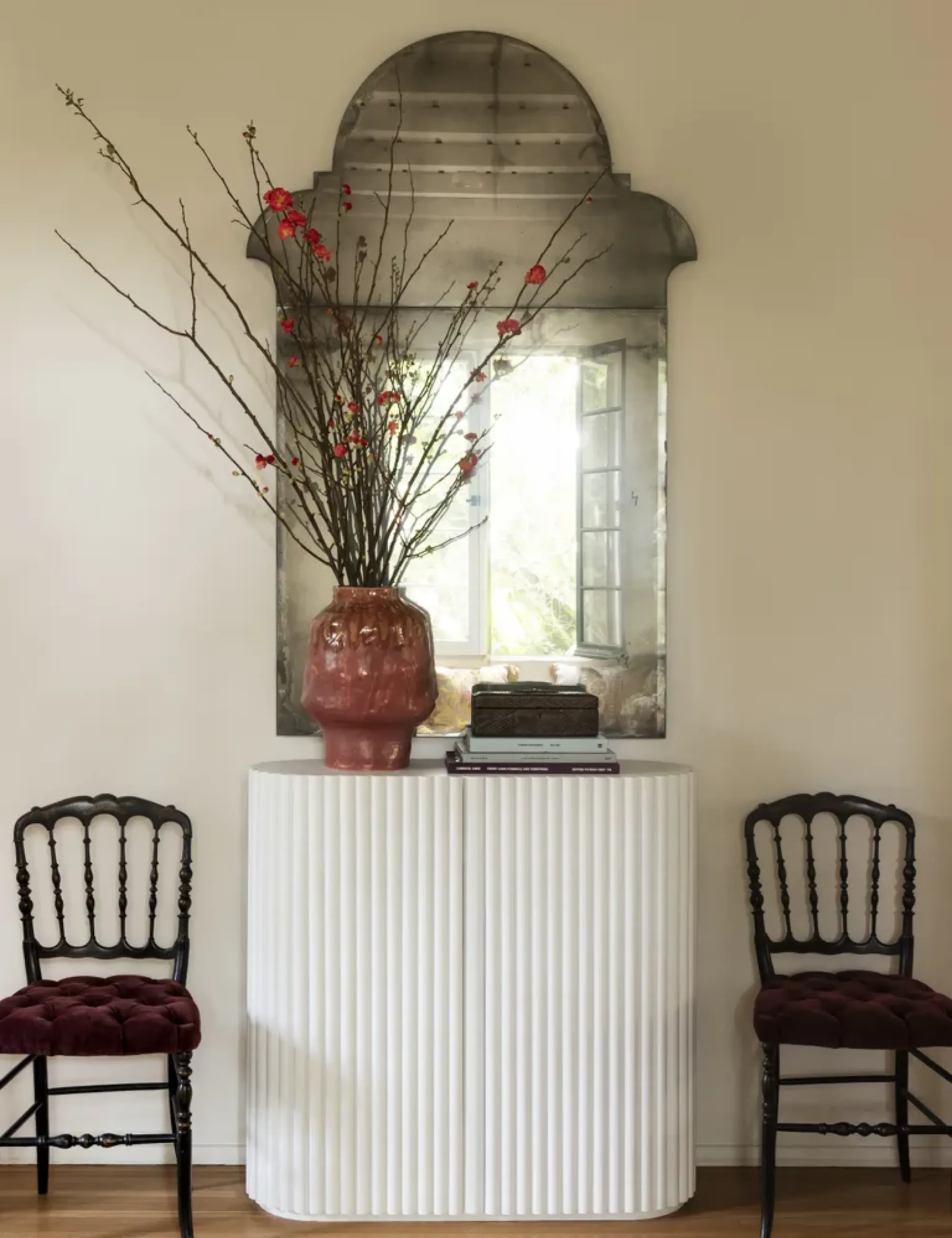 Starožitné zrcadlo visí nad bílou konzolou se dvěma polstrovanými jídelními židlemi lemujícími jednotku.' loading='lazy' title=