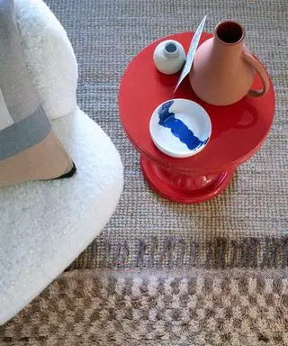 Zbliżenie na brązowy i beżowy dywanik w kratkę na beżowym dywanie z teksturą, prawy czerwony okrągły stolik boczny, ozdoby na stole, róg kremowego fotela z poduszką