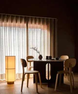 En svakt opplyst spisestue med en stor lysende stående lampe og gulv-til-tak-vinduer dekket av gardiner