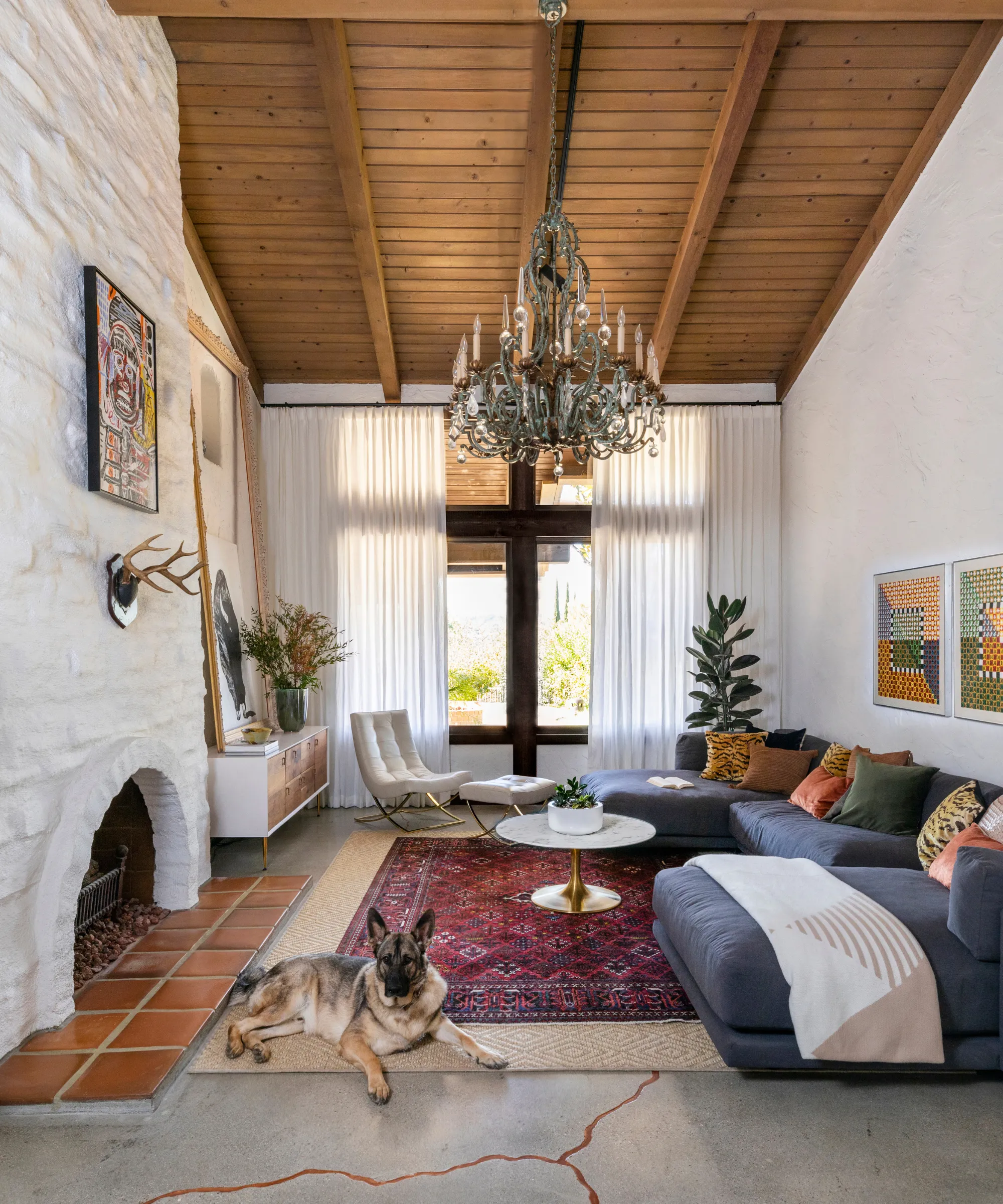 アーチ型の木製パネル天井のあるリビングルーム 枕がいくつかある大きなブルー/グレーのソファ 白い壁 背景に白いカーテンで覆われた床から天井までの窓 天井から吊り下げられた大きなシャンデリア、床のジュート絨毯の上にペルシャ絨毯が敷かれている' loading='lazy' title=