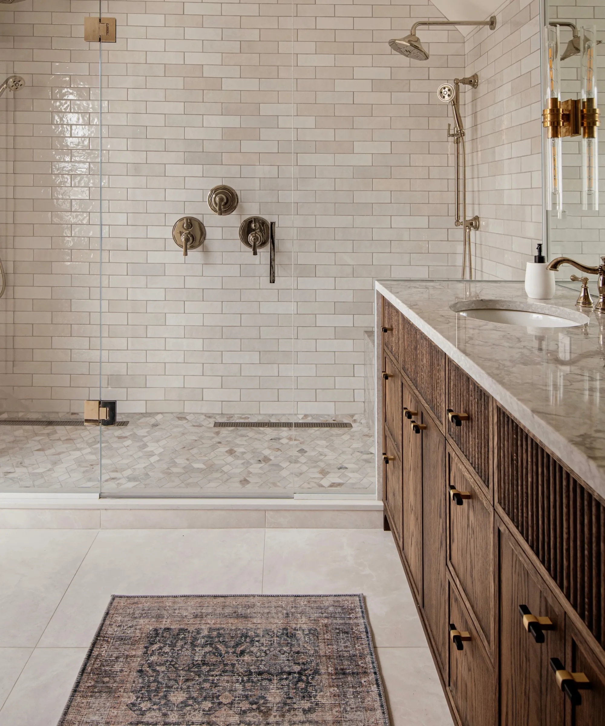 バスルームのシャワーにはオフホワイトのタイル張りの壁、化粧台用のベージュの大理石のカウンタートップ、その下にダークウッドのキャビネットがあり、床にはペルシャランナーラグが敷かれています。' loading='lazy' title=