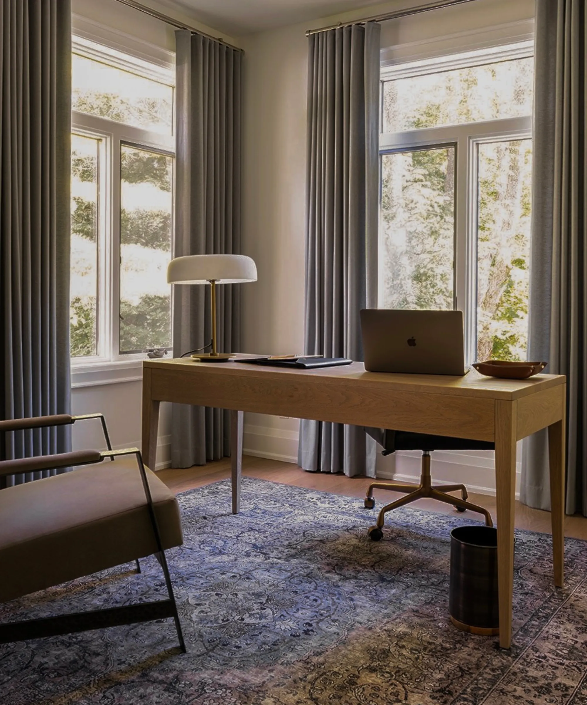 シンプルなホームオフィスには、床から天井まで覆われた大きな窓があり、ドレープがかかっているシンプルな木製の机にランプ、ラップトップと書類が置かれ、アクセントチェア、小さなゴミ箱、床にはペルシャ絨毯が敷かれています。' loading='lazy' title=
