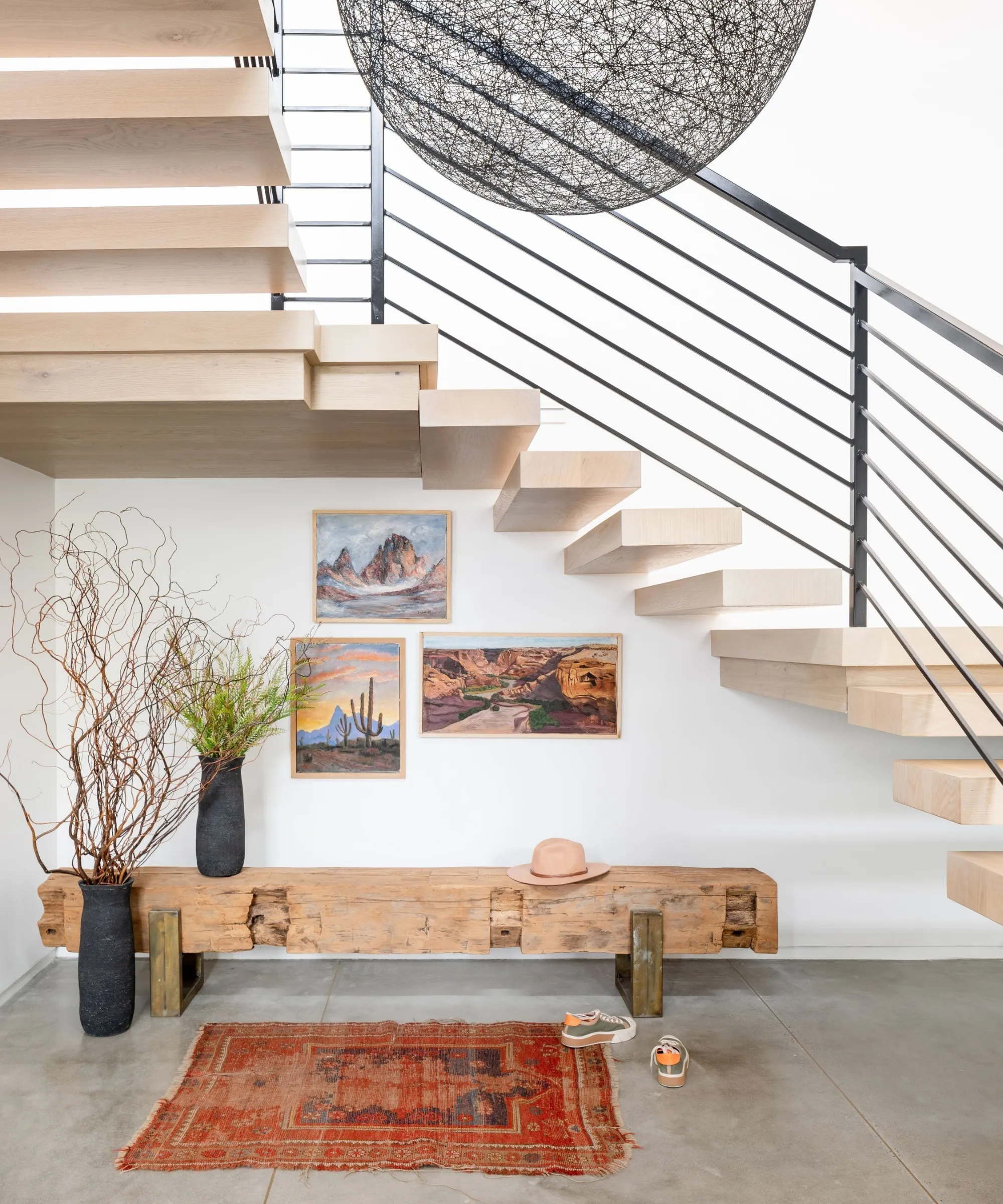 階段の下にある玄関のリビングエリア、傷んだ木製のベンチ、小さな傷んだペルシャ絨毯、植物が入った 2 つの花瓶、壁に掛けられたアートワーク' loading='lazy' title=