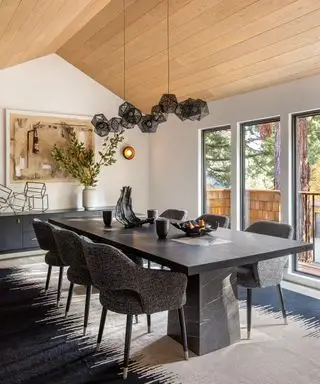 Großes Esszimmer mit Holzdeckenverkleidung, großem rechteckigen Marmor-Esstisch mit sechs gepolsterten Stühlen, schwarzem und cremefarbenem Teppich, schwarzer Metall-Cluster-Pendelleuchte, schwarzem niedrigem Sideboard mit gerahmtem Kunstwerk und Blumenvase