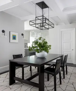 Grau gestrichenes Esszimmer mit weißer Kassettendeckenverkleidung, schwarzem Esstisch und vier schwarzen Esszimmerstühlen, geometrischer Hängeleuchte aus schwarzem Metall, Pflanze in weißer Vase auf dem Tisch, Wandkunst und Wandlampen an der Wand, schwarzer und cremefarbener Teppich im geometrischen Stil