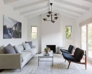 Weiß gestrichenes Wohnzimmer mit weißer Holztäfelung und Balken, großem gestreiften Teppich, cremefarbenem Sofa und zwei Loungesesseln, weißem und schwarzem Couchtisch, Hängelampe aus Metall und Berglandschaftskunstwerk