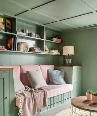 Sitzbereich mit grün gestrichenen Wänden und Decke, Decke mit Täfelung, Sitzbereich aus Holzvertäfelung, ausgestattet mit rosa Kissen und Accessoires, natürlicher Rundteppich und Couchtisch, Regale mit dekorativen Accessoires und Büchern