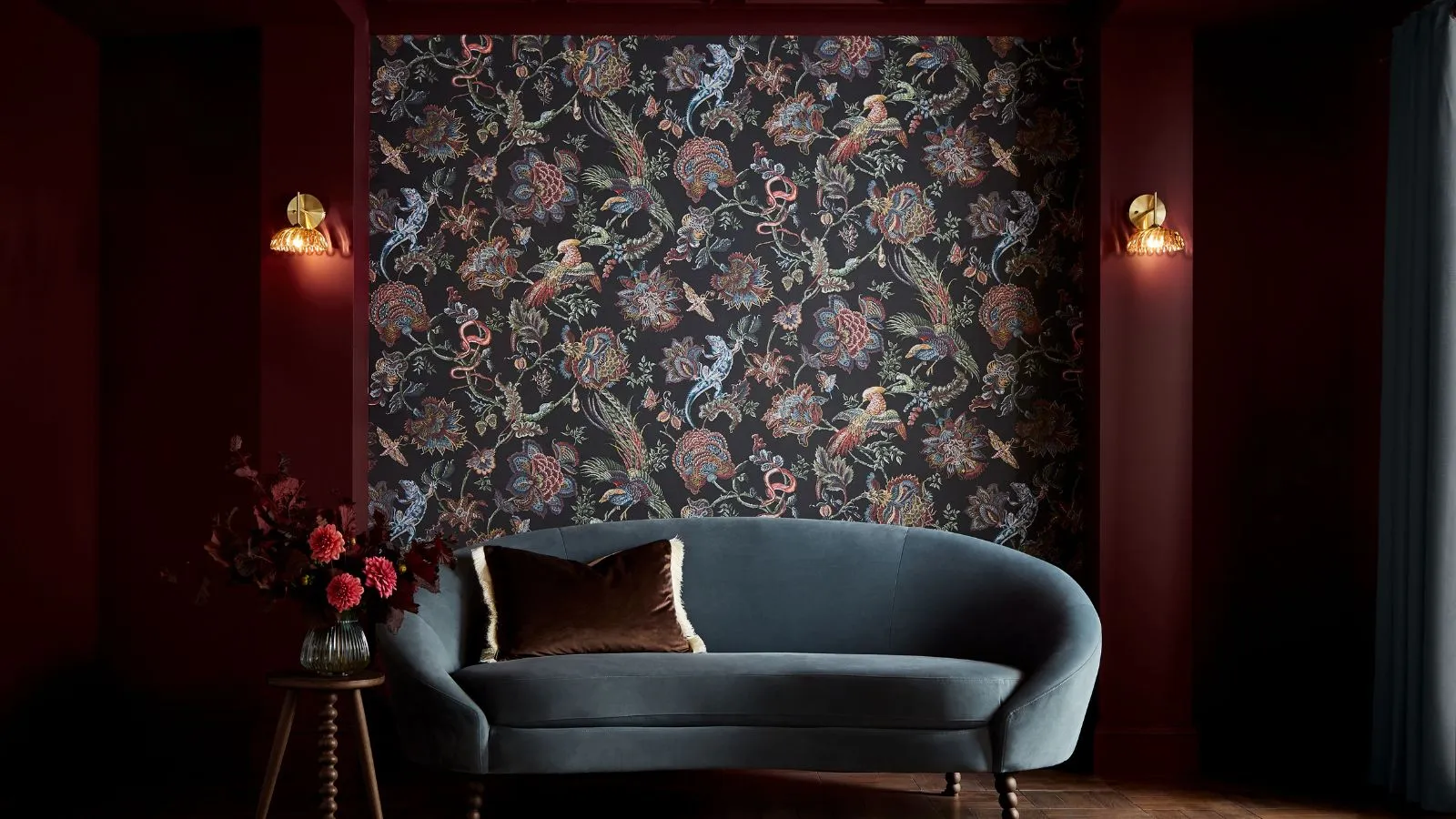 Salon peint en rouge foncé avec appliques murales en papier peint et canapé incurvé en velours bleu avec coussin et table d'appoint avec vase contenant des roses' loading='lazy' title=