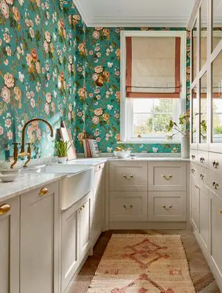 Cocina de galera gris con papel tapiz verde azulado con flores rosadas. Ventana enmarcada en blanco en la pared del fondo con persiana de lino blanco y rojizo
