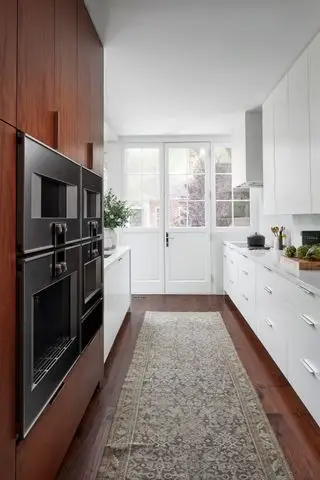 cozinha galley com armários de nogueira e armários brancos com forno duplo e tapete oriental sobre piso de madeira escura