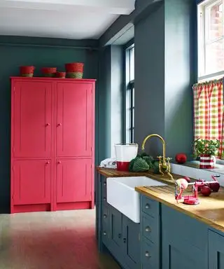 Cozinha pintada em azul escuro com armário independente de madeira vermelho brilhante, piso de madeira clara, pia com torneira de latão, meias cortinas de algodão multicoloridas