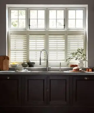 Grande janela acima da pia com veneziana branca, armários de cozinha marrom escuro, pia de cozinha, bancadas decoradas com utensílios de cozinha e alimentos