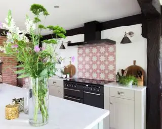 Et hvitt kjøkken med en rosa mønstret flisplate, svart stekeovn og stor vase med blomster i forgrunnen.
