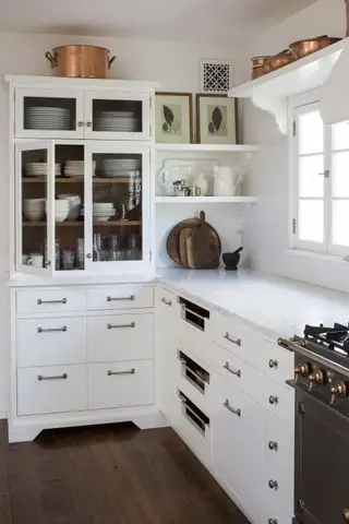 Weiße Küche mit weißen Schränken, Marmorarbeitsplatten, Schrank mit Glasfront, offenen Regalen, dunklem Holzboden, Kupferpfannen und Kunstwerken