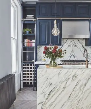 Un'isola da cucina rivestita in marmo bianco, con mobili blu sul resto della cucina, portabottiglie e scala per raggiungere gli scaffali più alti