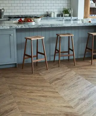Küche mit Vinylboden aus dunklem Holz, blau gestrichenen Schränken und Kücheninsel, Hockern aus dunklem Holz, Arbeitsplatte aus grauem Marmor und weißen Wandfliesen