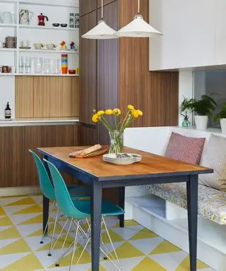 Wohnküche mit gelb-weißem, geometrischem Vinylboden, dunklen Holzschränken und Esstisch, Sitzbänken mit Kissen, blauen Esszimmerstühlen, niedrig hängenden Hängelampen